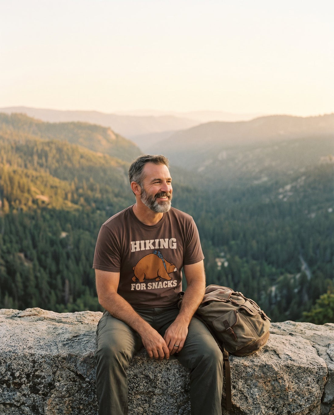 Bearded hiker resting on a granite ledge above a forested valley at golden hour, wearing the Hiking for Snacks tee in Espresso
