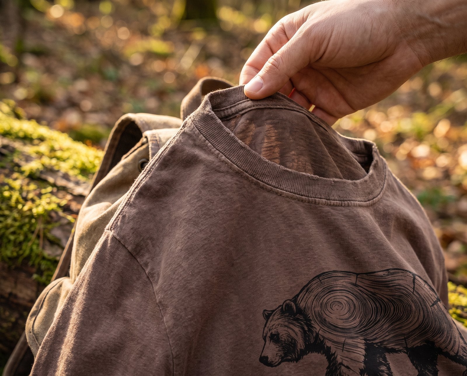 Hand lifting the collar of a soft espresso garment-dyed tee draped over a canvas daypack on a mossy log
